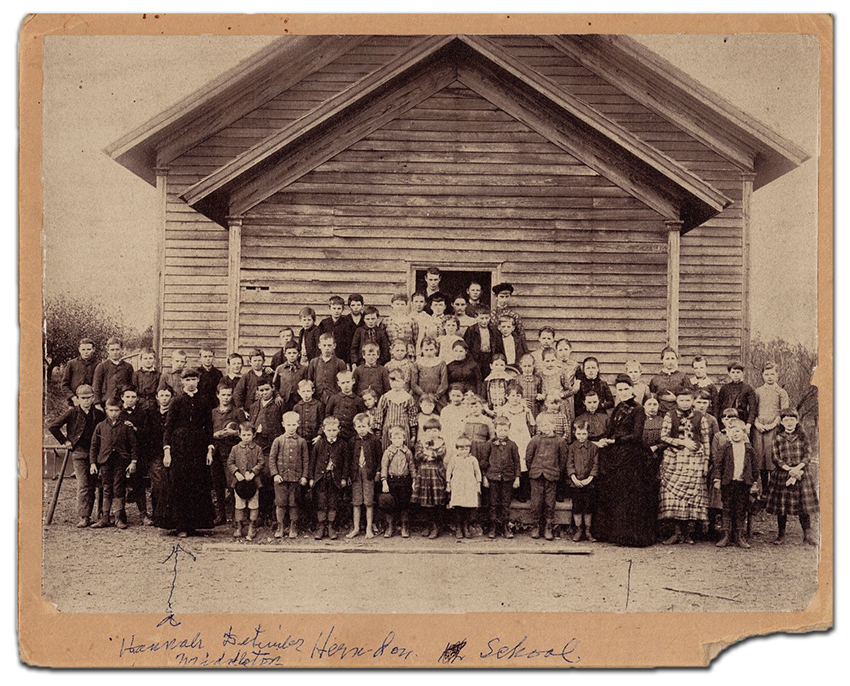 Black and white photograph of the first Herndon public school. Two teachers and approximately 70 children are pictured in front of a single-room, single story frame school building.  