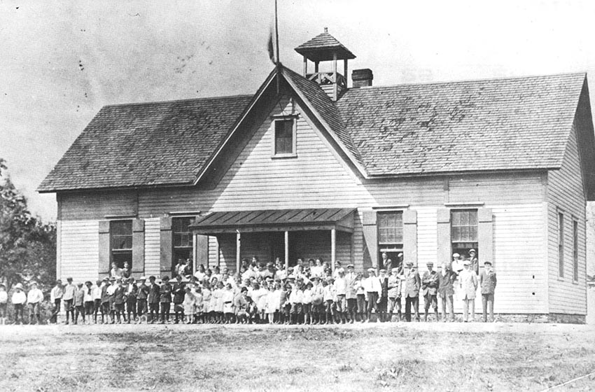 Black and white photograph of the three-room Herndon School. The building has been enlarged substantially. A large group of students is pictures standing in front of the school.